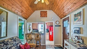 Un accogliente soggiorno con soffitto in legno, un divano decorato e un'area pranzo con sedie. Si intravede una cucina con elettrodomestici rossi attraverso una porta aperta.