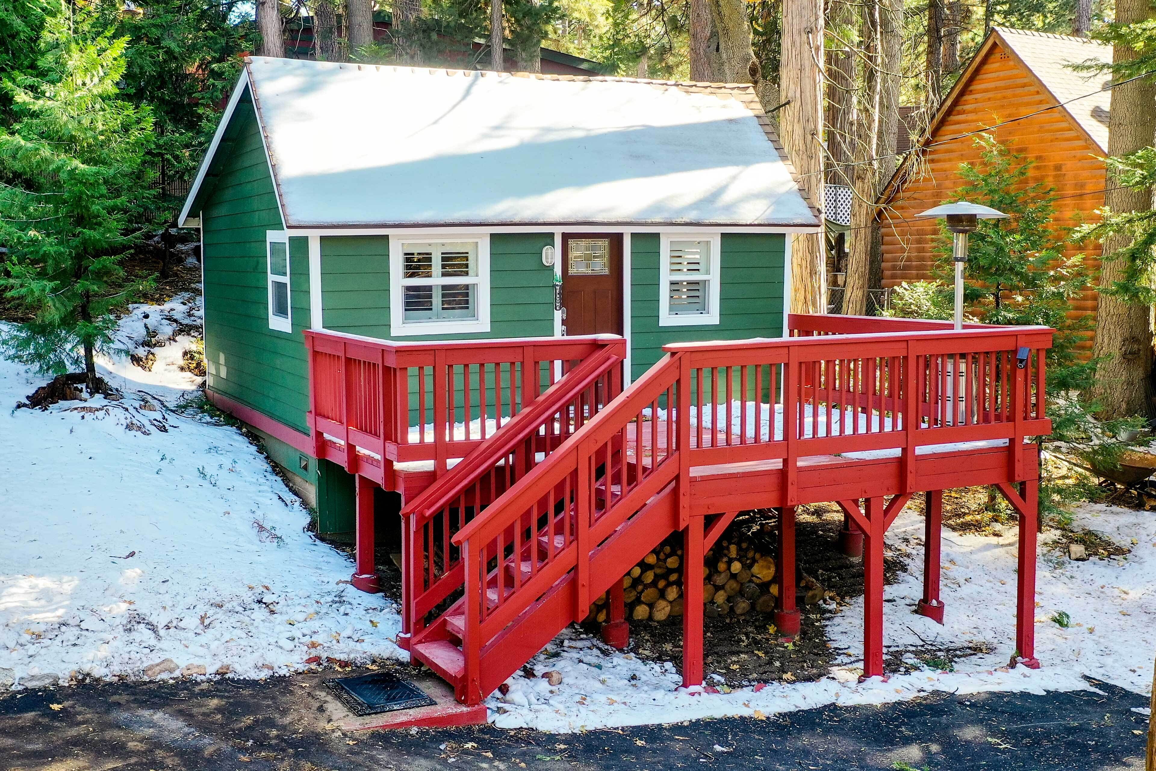 A green cabin with a red deck and stairs, surrounded by trees and snow.