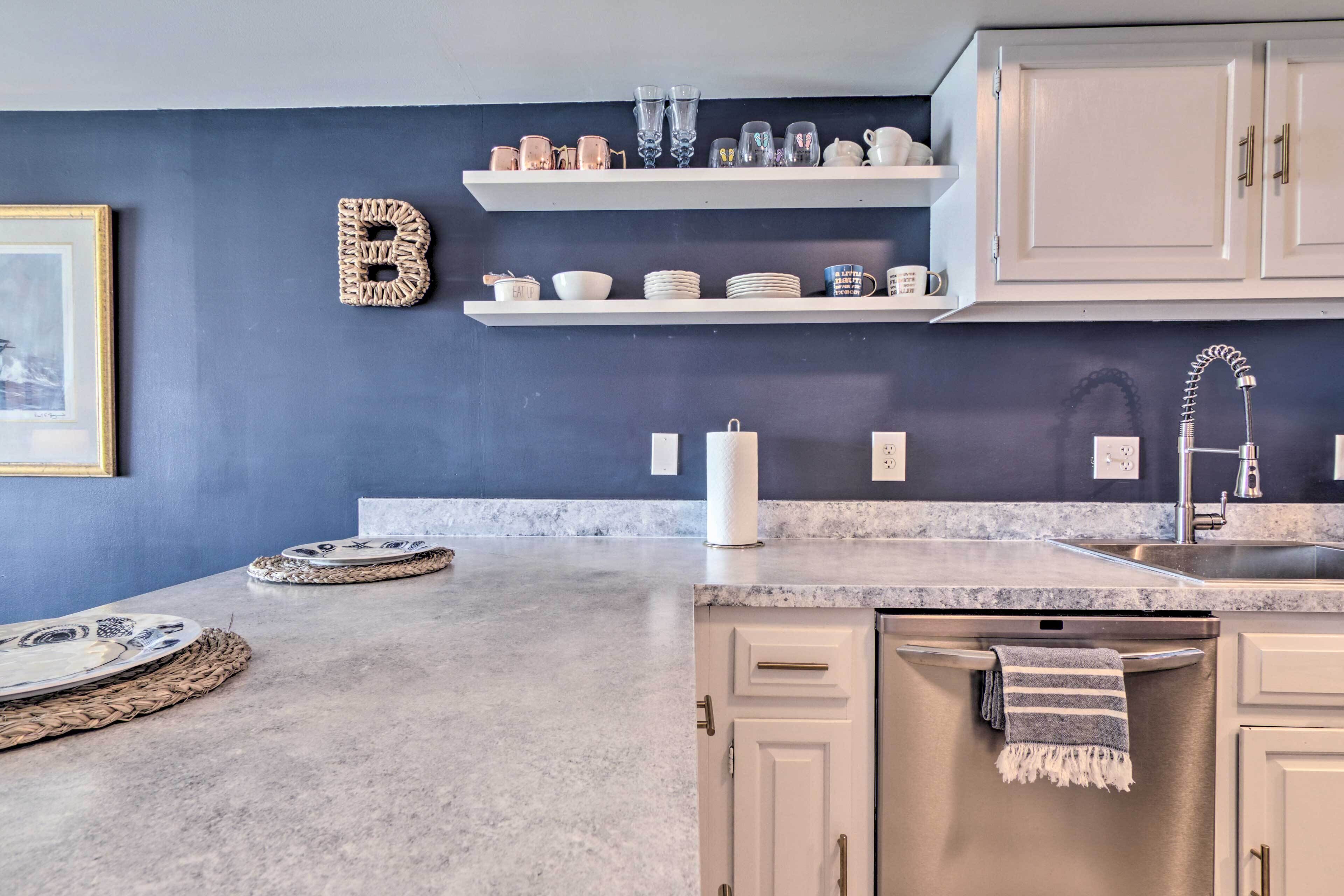 Een moderne keuken met witte kasten, een granieten aanrechtblad en een spatwand. Boven het aanrecht staan planken met servies en glazen.