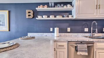Een moderne keuken met witte kasten, een granieten aanrechtblad en een spatwand. Boven het aanrecht staan planken met servies en glazen.