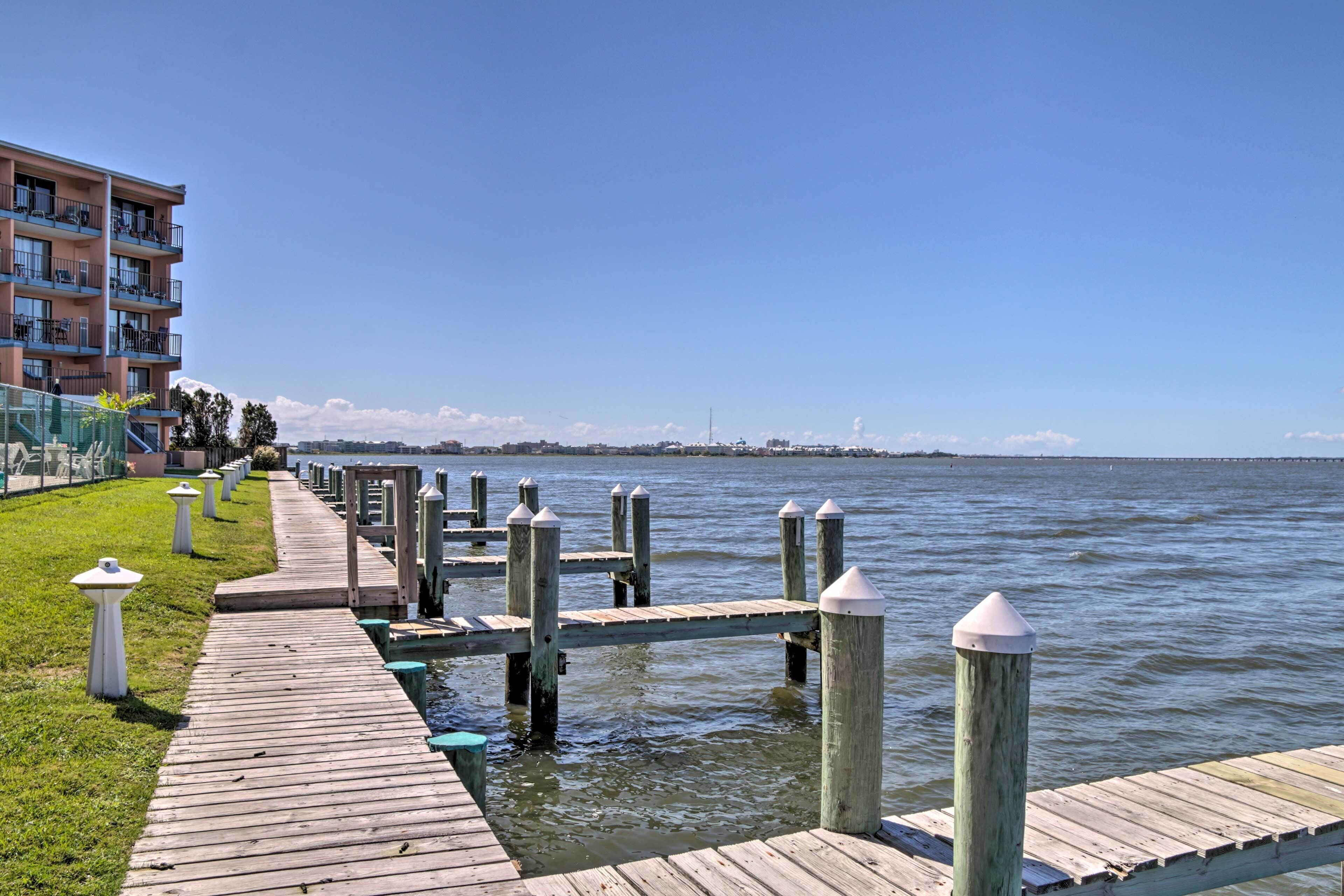 A wooden pier extending into calm water, with a grassy area and a multi-story building on the left.