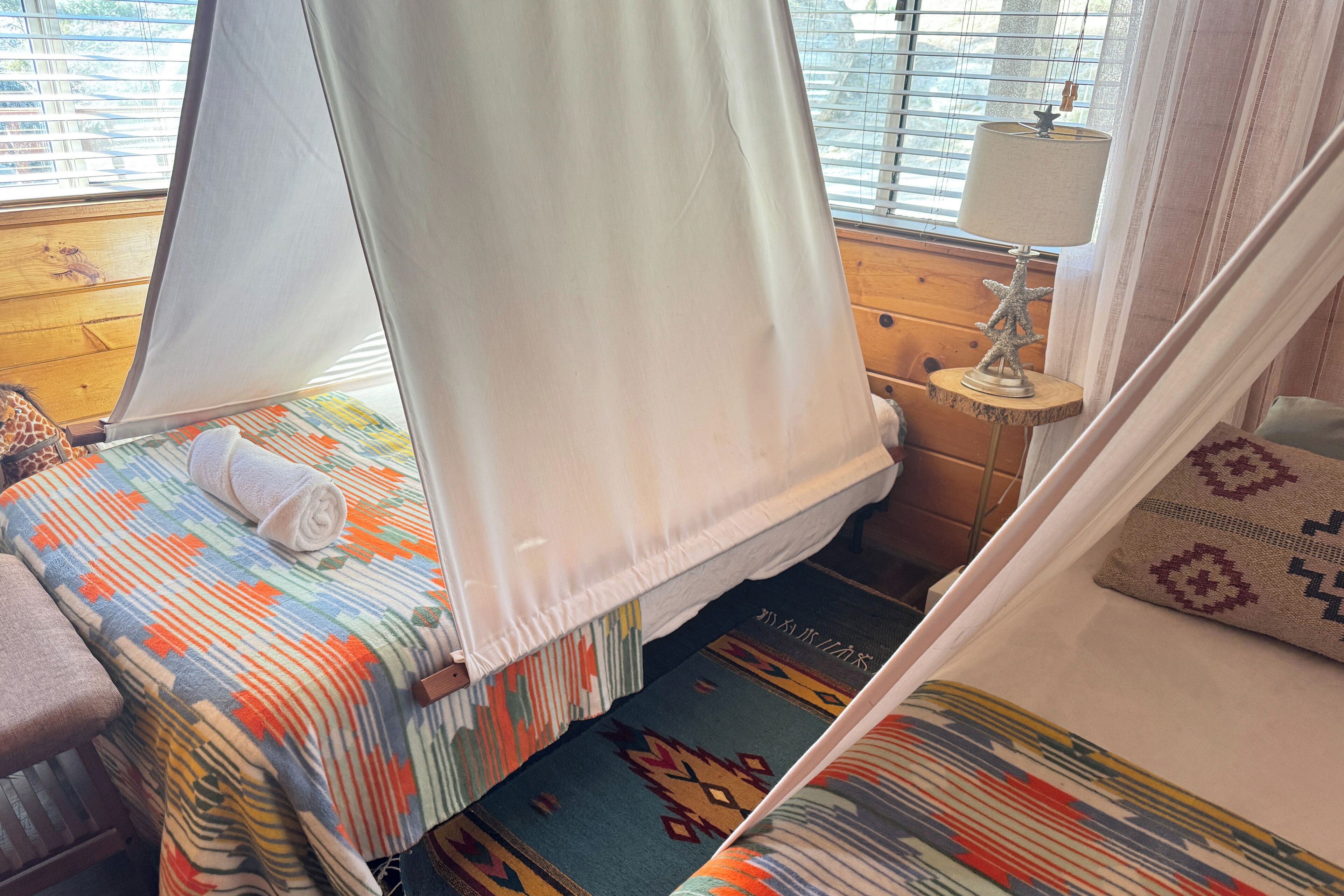 Een kamer met een bed, een raam met zonwering, een lamp en een gordijn.