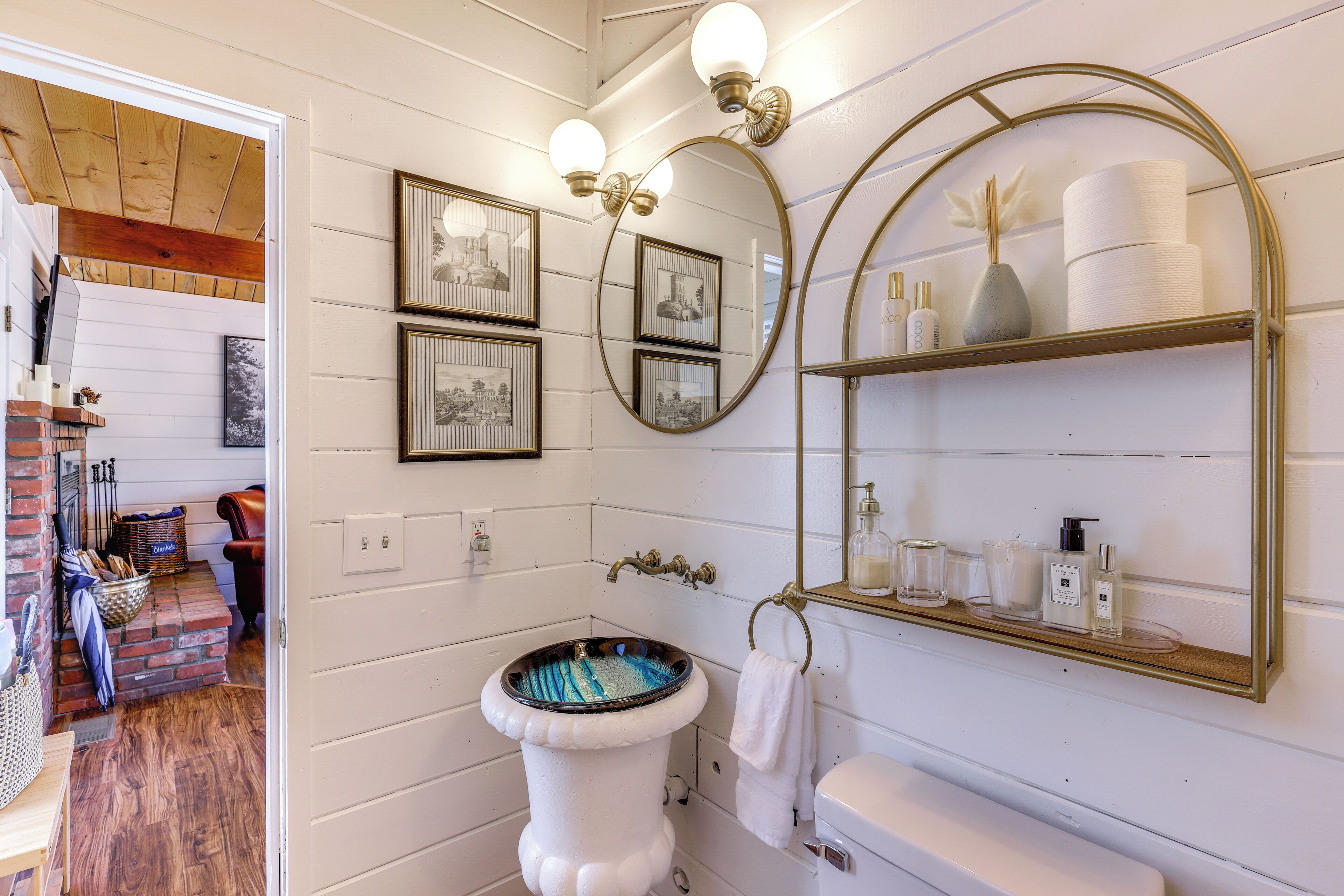 A bathroom with a white pedestal sink, a mirror with a gold frame, and a shelf with toiletries.
