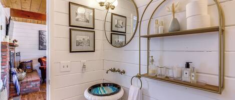 A bathroom with a white pedestal sink, a mirror with a gold frame, and a shelf with toiletries.