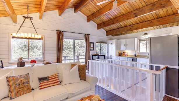 Uma sala de estar ampla com um sofá branco, vigas de madeira no teto e uma cozinha com armários brancos e uma geladeira de aço inoxidável.