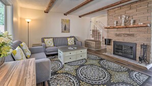Un salon avec un canapé gris, un tapis à motifs, une table basse en bois et une cheminée avec un mur en briques.