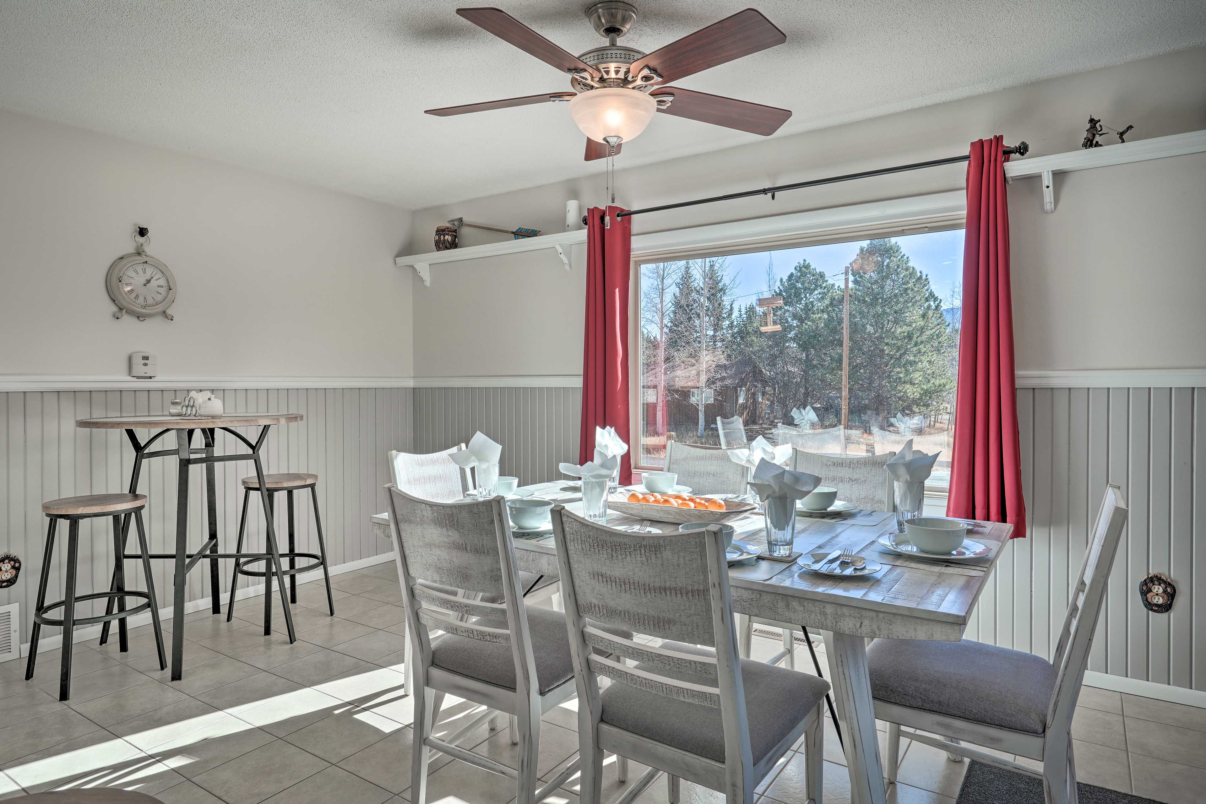 Un coin repas avec une table dressée pour quatre personnes, deux tabourets de bar, un ventilateur de plafond et une fenêtre ornée de rideaux rouges.