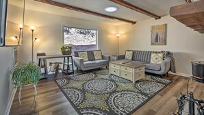 Un salon avec un canapé gris, une table basse et un tapis à motifs. Il y a une fenêtre donnant sur des arbres et une cheminée.