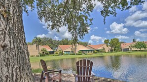 A serene outdoor seating area with two Adirondack chairs by a pond, surrounded by a well-maintained lawn and a row of houses with red roofs. - Luxury Naples Villa - Walk to Vanderbilt Beach! (Naples)