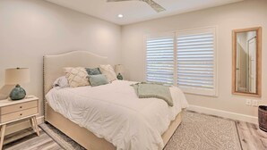 A bedroom with a large bed, a nightstand, a lamp, a ceiling fan, a window with blinds, and a mirror on the wall. - Luxury Naples Villa - Walk to Vanderbilt Beach! (Naples)