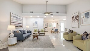 A spacious living room with a grey sofa, yellow armchairs, and a coffee table. There are two side tables with lamps, a ceiling fan, and artwork on the walls. - 1 Mi to Dtwn Palm Springs: Home w/ Fire Pit! (Palm Springs)