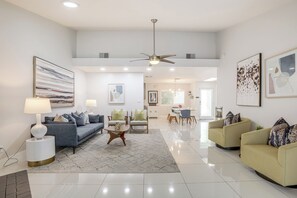 A spacious living room with a grey sofa, yellow armchairs, and a coffee table. There are two side tables with lamps, a ceiling fan, and artwork on the walls. - 1 Mi to Dtwn Palm Springs: Home w/ Fire Pit! (Palm Springs)