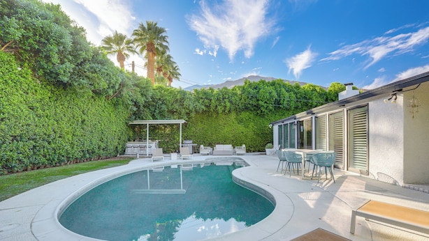 A swimming pool with a curved edge, surrounded by a white deck. There are outdoor furniture pieces and a pergola. - 1 Mi to Dtwn Palm Springs: Home w/ Fire Pit! (Palm Springs)