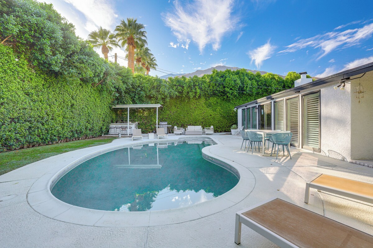 A swimming pool with a curved edge, surrounded by a white deck. There are outdoor furniture pieces and a pergola.