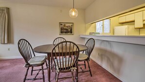 Een eetgedeelte met een ronde tafel en vier houten stoelen, een hanglamp, een lijst met een afbeelding aan de muur, en een keukenblad met kasten.