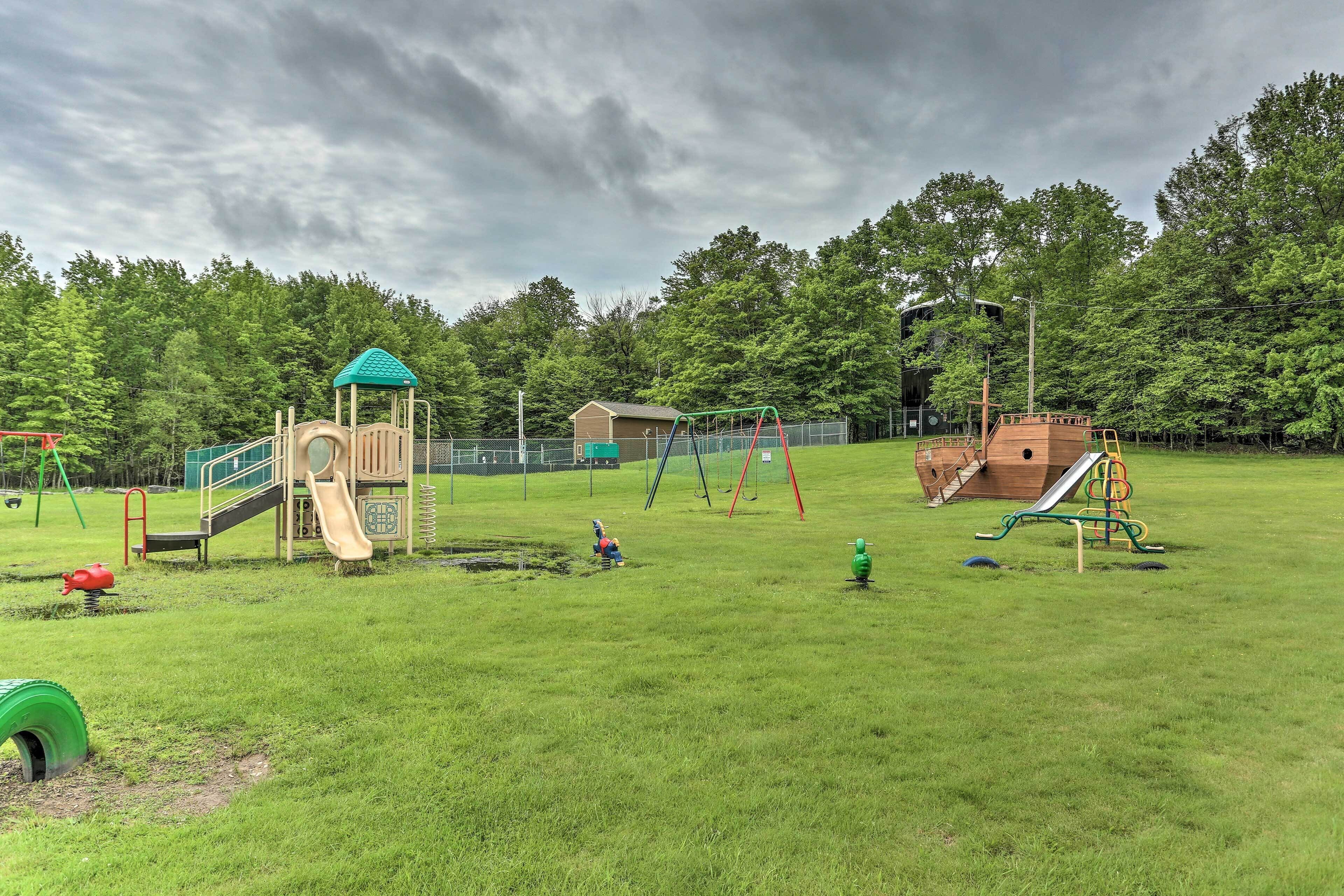A playground with various play structures, including a slide and swings, surrounded by a grassy field and trees.