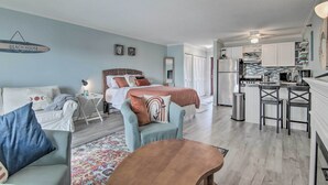 A modern bedroom with a bed, bedside tables, and a nightstand. There is a kitchenette with a refrigerator, stove, and sink. The room has light blue walls and a wooden floor. - Central Studio w/ Deck: Walk to Shops & Dining! (Ocean City)