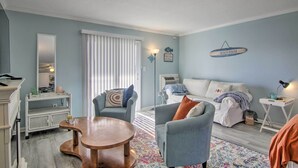 A living room with a sofa, armchairs, a coffee table, and a dining table. There is a window with blinds, a wall-mounted sign, and a lamp on a side table. - Central Studio w/ Deck: Walk to Shops & Dining! (Ocean City)