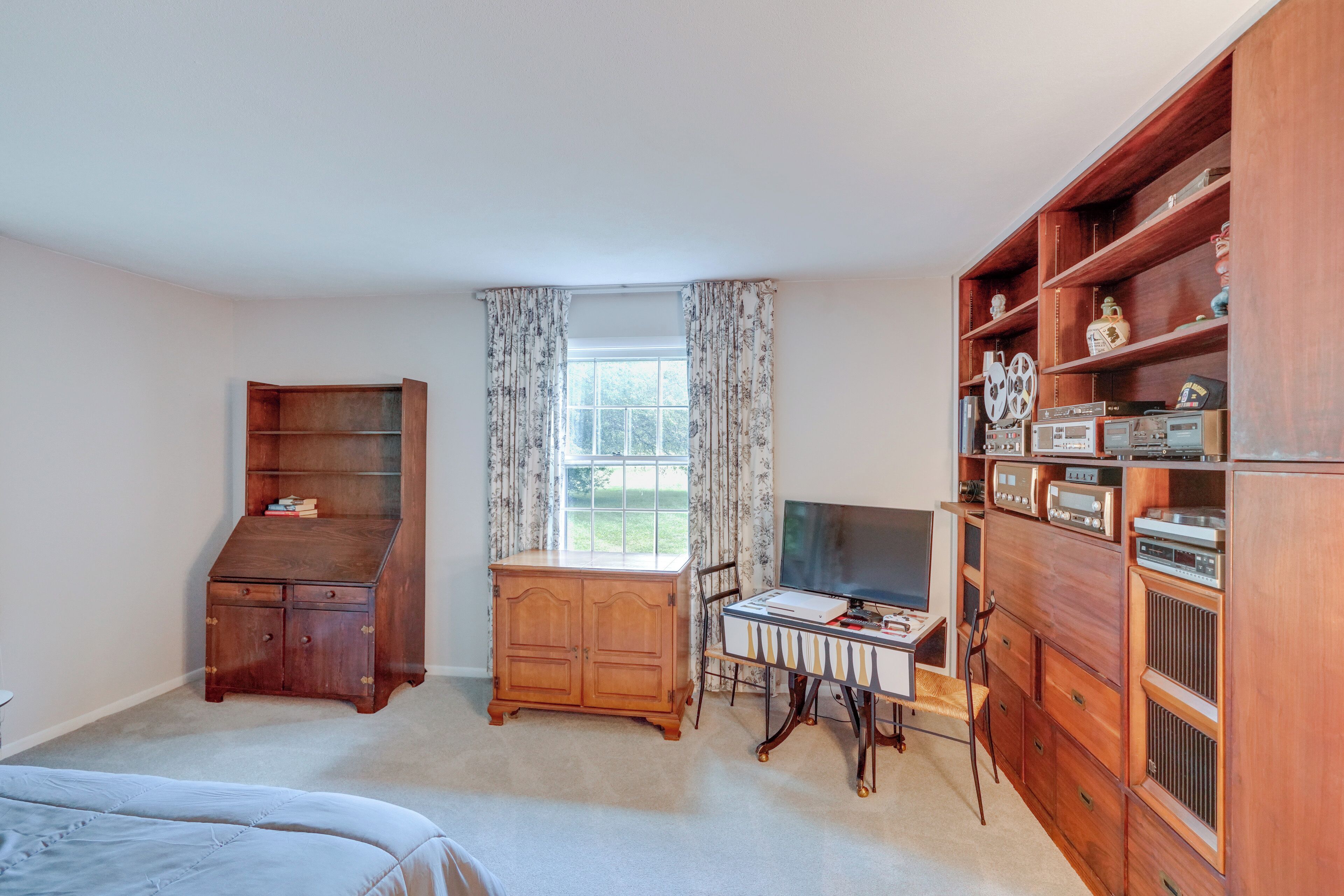 Een slaapkamer met een bed, een houten dressoir, een bureau met een computer en een grote houten kast met diverse spullen.