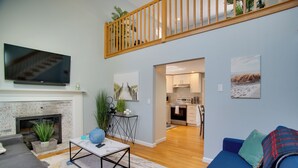 A modern living room with a fireplace, a flat-screen TV, a dining area, and a kitchen. - Cape Cod Family Home w/ Yard: Walk To Beach! (West Yarmouth)