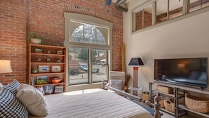 Une chambre moderne avec un grand lit, une bibliothèque en bois, un téléviseur et une vue sur un bâtiment en briques, visible à travers une grande fenêtre.