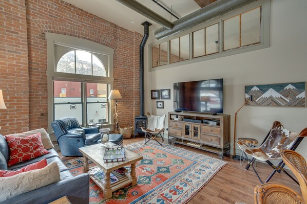 A cozy living room with a brick wall, a large window, a fireplace, and a flat-screen TV. - Updated Rustic-chic Condo on Ouray's Main Street! (Ouray)