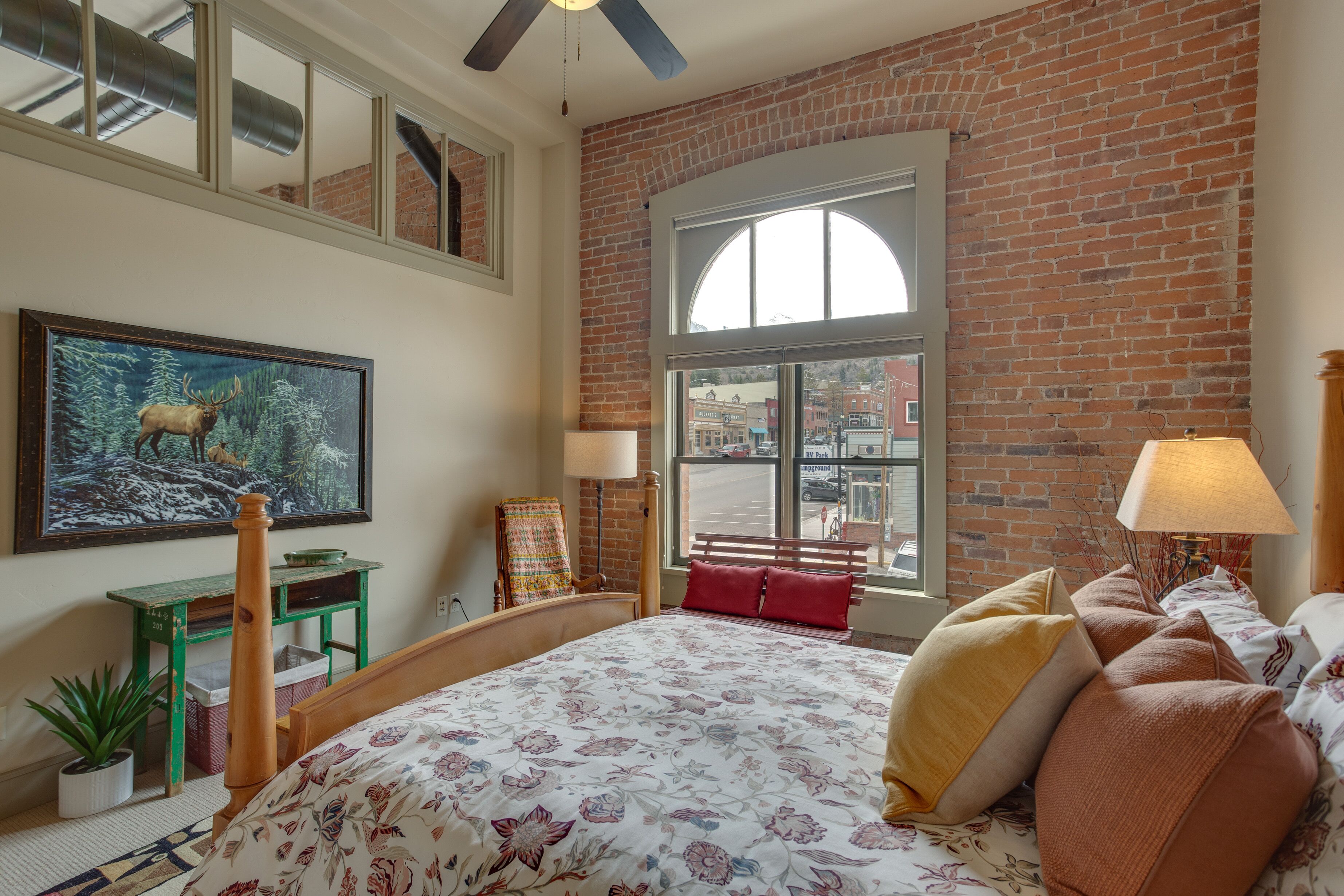Ein Schlafzimmer mit einem großen Bett, einem grünen Schreibtisch, einem Stuhl, einem Fenster mit Blick auf die Stadt und einem Elchgemälde an der Wand.