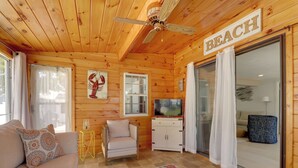 A cozy cabin interior with a wooden ceiling, a ceiling fan, a sofa with patterned cushions, a small armchair, a television, and a large sliding glass door labeled "BEACH". - Charming West Yarmouth Home ~ 2 Mi to Beach! (West Yarmouth)