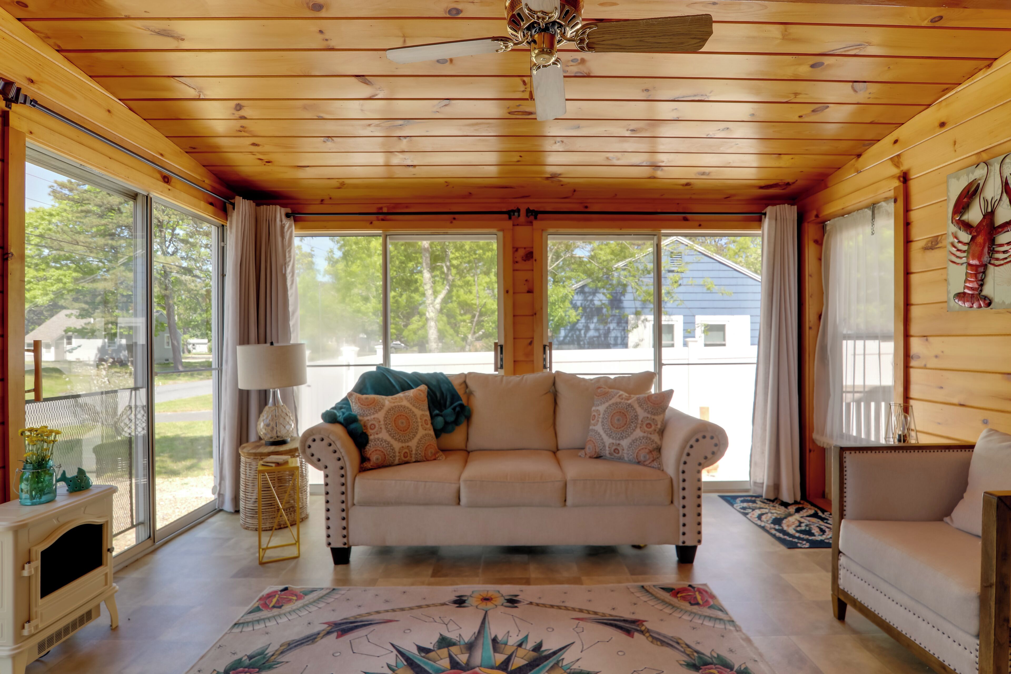 Une pièce chaleureuse avec un plafond en bois, un canapé beige, un tapis à motifs et de grandes fenêtres donnant sur l’extérieur.