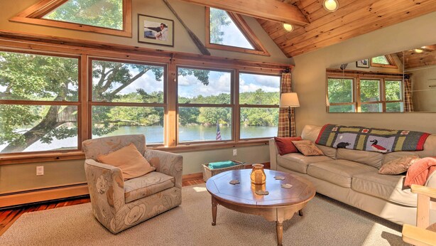 Una sala de estar acogedora con un gran ventanal con vistas a un lago, un sofá, un sillón y una mesita de centro.