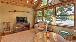 Un salón acogedor con televisor, muebles de madera, ventanas amplias y vista a árboles.