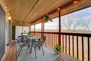 An outdoor patio with a wooden deck, a table with chairs, hanging plants, and a view of trees and hills.
