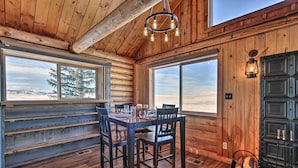 A wooden cabin interior with a dining table and chairs, large windows with a view of the mountains, and a fireplace with a lantern.