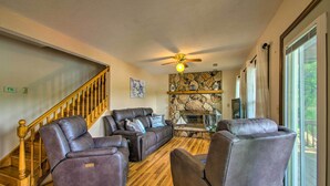 A spacious living room with a stone fireplace, leather recliners, a wooden staircase, and large windows.