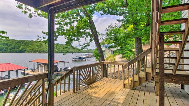A wooden deck with a railing overlooking a lake, docked boats, and a covered area with tables and chairs.