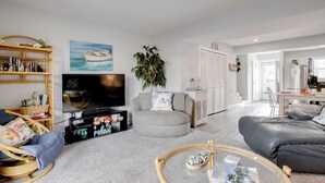 Una sala de estar con televisor, un sofá gris, una silla de mimbre, una mesita redonda y una estantería.