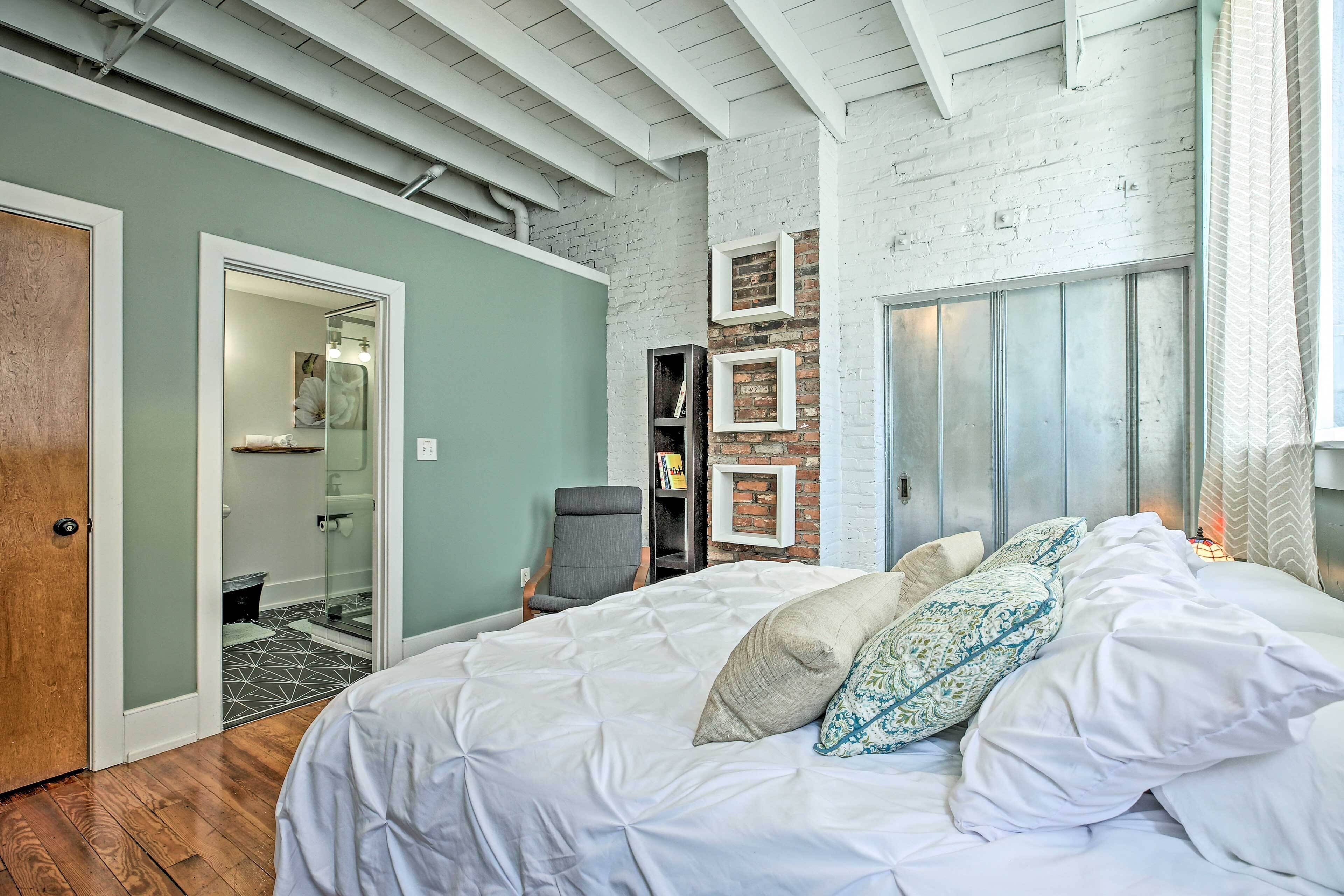 A bedroom with a large bed, a chair, a wardrobe, and a brick wall.