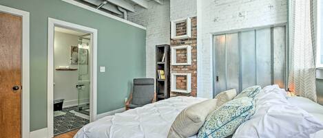 A bedroom with a large bed, a chair, a wardrobe, and a brick wall.