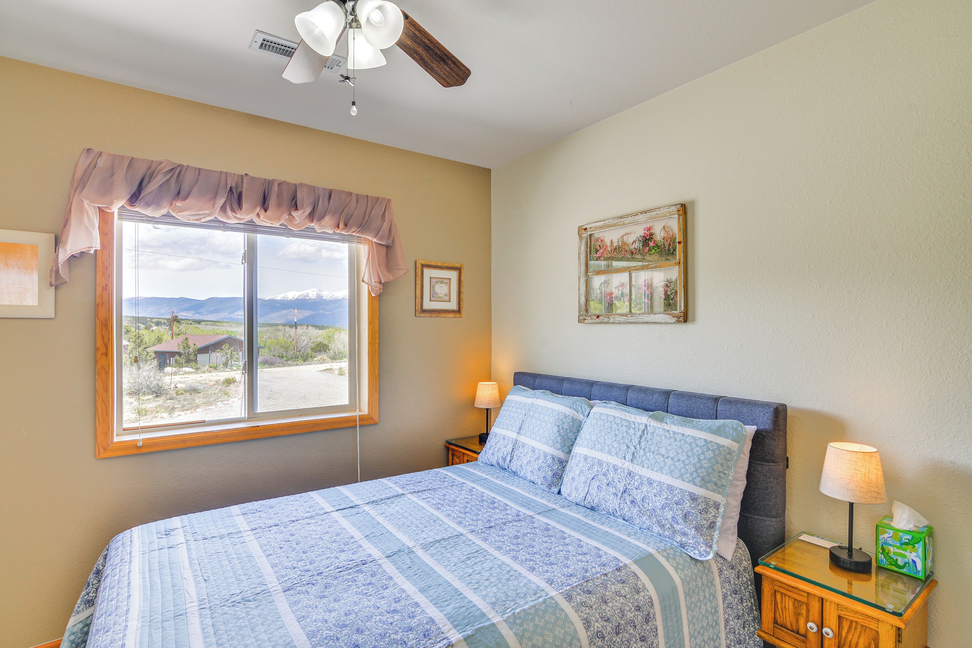 A bedroom with a large bed, a window with a view of the mountains, a ceiling fan, and a bedside table with a lamp and tissues.