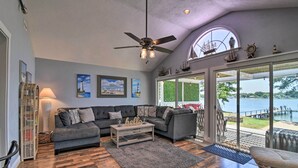 A spacious living room with a large gray sectional sofa, a wooden coffee table, and a ceiling fan. There are nautical decorations on the wall and a view of a lake through large glass doors. - Riverfront Cottage: Fire Pit & Kayaks (Deltaville)