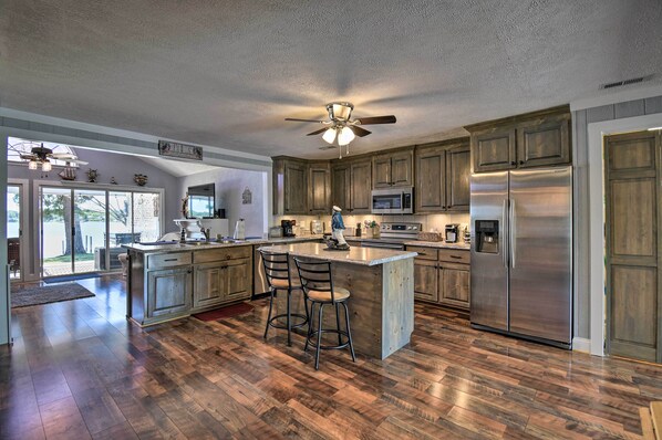 A spacious kitchen with wooden cabinets, a large island with bar stools, stainless steel appliances, and a ceiling fan. - Riverfront Cottage: Fire Pit & Kayaks (Deltaville)