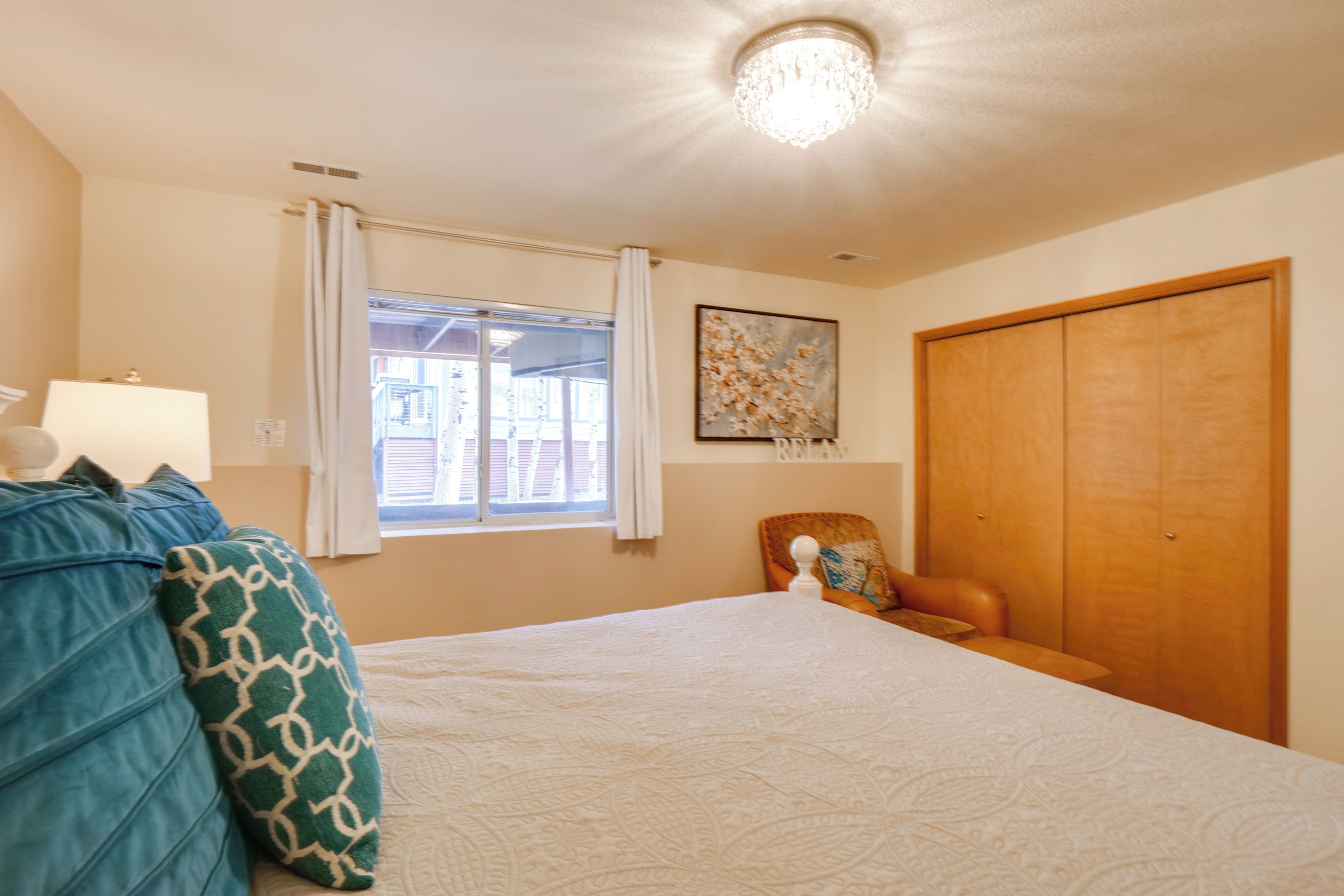 Un dormitorio con cama, una silla, un armario, una ventana con cortinas y una lámpara en el techo.