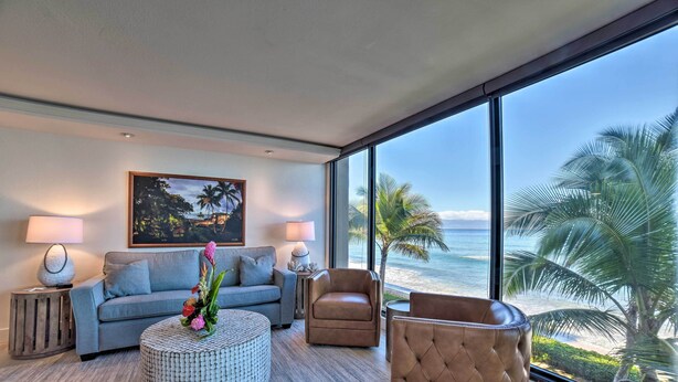 A modern living room with a large window overlooking a beach, a gray sofa, a round coffee table with flowers, and a brown armchair. - Whale & Sunset Views: Oceanfront Lahaina Condo (Kaanapali)