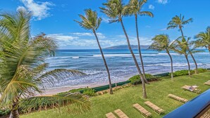 A beach view with palm trees, a grassy area with lounge chairs, and a clear blue sky. - Whale & Sunset Views: Oceanfront Lahaina Condo (Kaanapali)