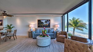 A modern living room with a round coffee table, a sofa, and a chair. There is a painting on the wall and a view of the beach through large windows. - Whale & Sunset Views: Oceanfront Lahaina Condo (Kaanapali)
