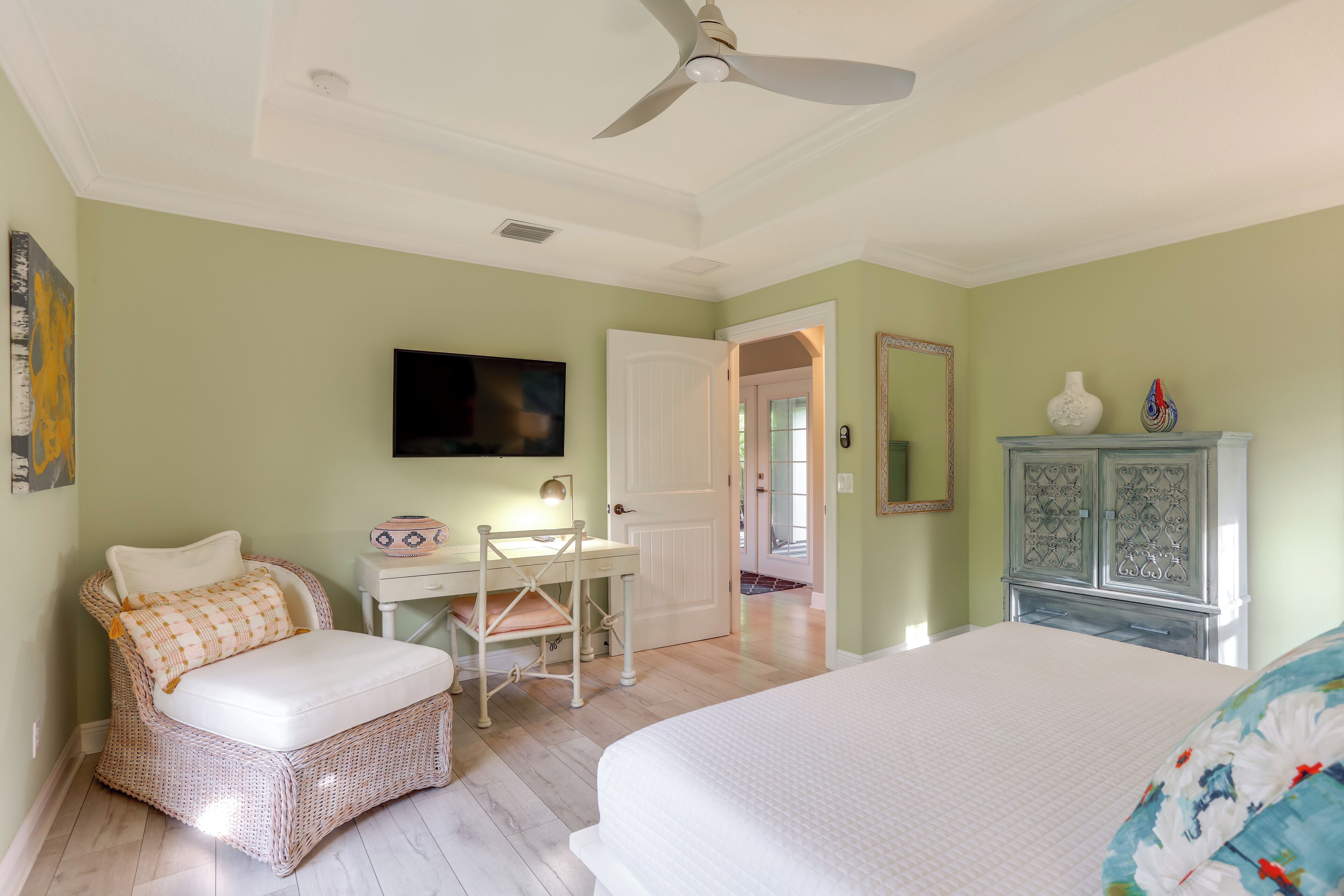 A bedroom with a bed, a wicker chair, a desk, a television, a mirror, and a dresser.