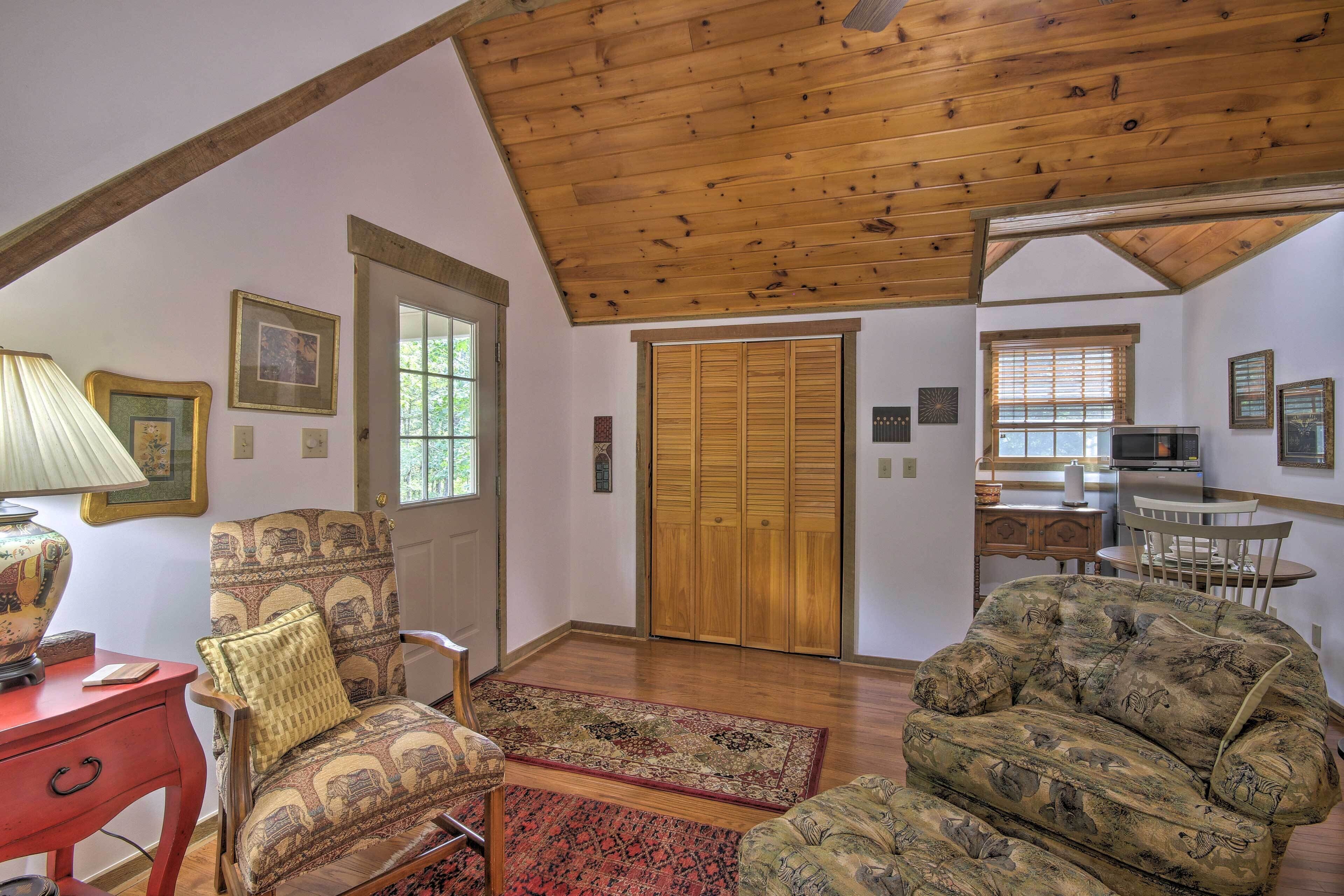 A spacious living room with a high ceiling, wooden floors, and a large window. There is a sofa, a chair, and a table with a lamp.