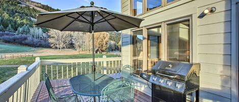 Een terras met een barbecue, buitentafel en stoelen, met uitzicht op een aangelegde tuin en een woning.