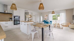 A modern kitchen with white cabinetry, a wooden countertop, and a dining area with white chairs and a wooden table. There are two hanging wicker lamps above the dining table. - Clair Jeanne - Climatisation - Plage (Notre-Dame-de-Monts)
