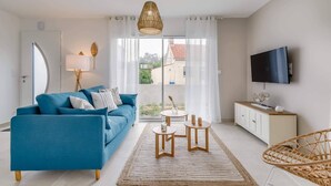A modern living room with a blue sofa, a wooden coffee table, a rattan chair, a TV, and a view of the outside. - Clair Jeanne - Climatisation - Plage (Notre-Dame-de-Monts)
